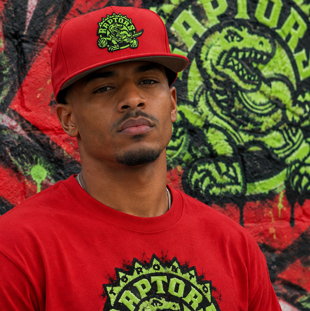 A red snapback cap featuring an embroidered green and black Toronto Raptors logo on the front, with a visible Mitchell & Ness sticker on the flat brim, set against a plain white background.
